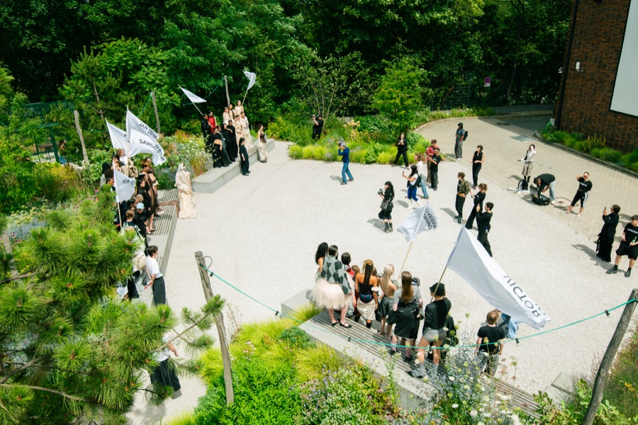 Aerial view of the LETTE SS27 garden runway at Atelier Gardens, models carrying white flags through the greenery