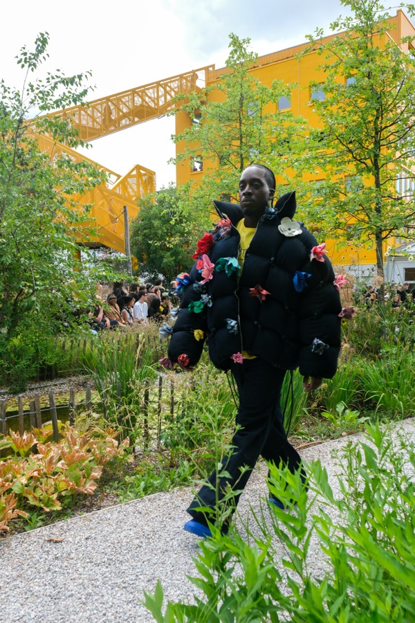 Model in a black puffer coat with fabric flowers walking the garden path, seated guests visible behind