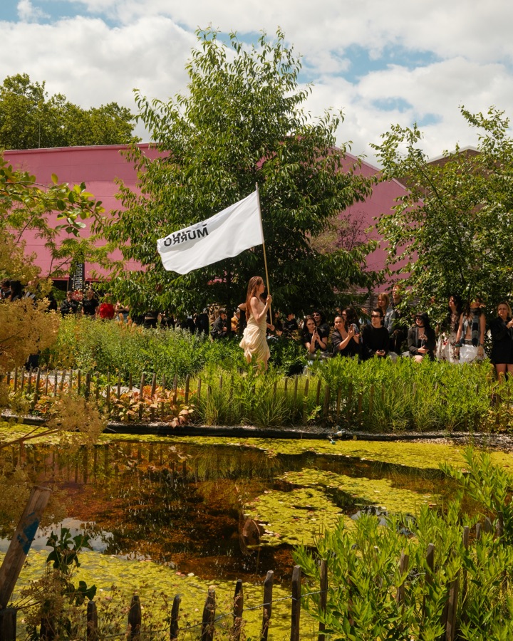 Model carrying a white flag along the garden runway beside a pond
